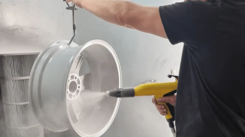 A painter spraying white paint onto an alloy wheel hanging in a spray booth